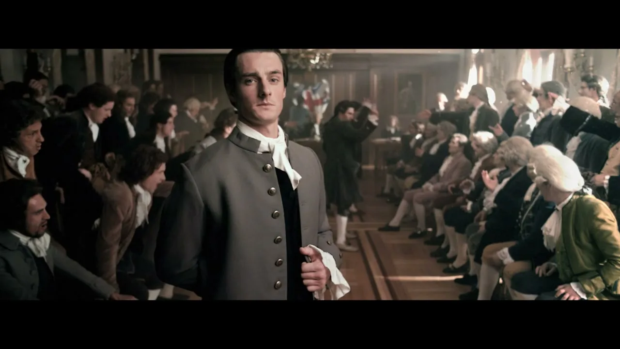 A man in 18th-century attire stands in a crowded assembly hall, looking directly at the camera as people around him react with agitation.