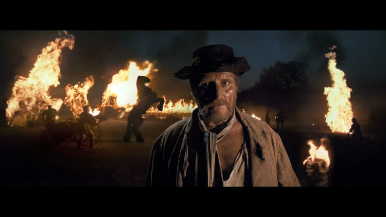 A rugged man in a hat and simple clothes stands in the foreground, looking directly at the camera, while a chaotic scene of fires and people unfolds in the background at night.