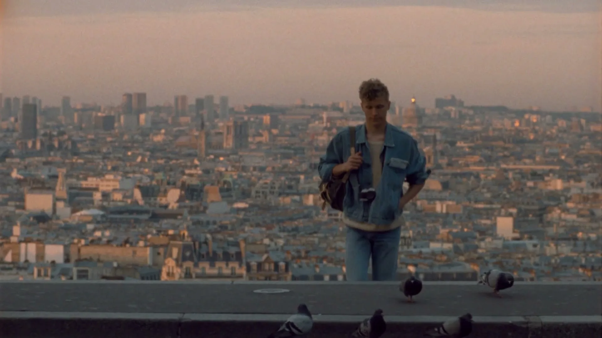 A young man in a denim jacket stands on a rooftop overlooking the city of Paris at dusk, with pigeons on the ground.