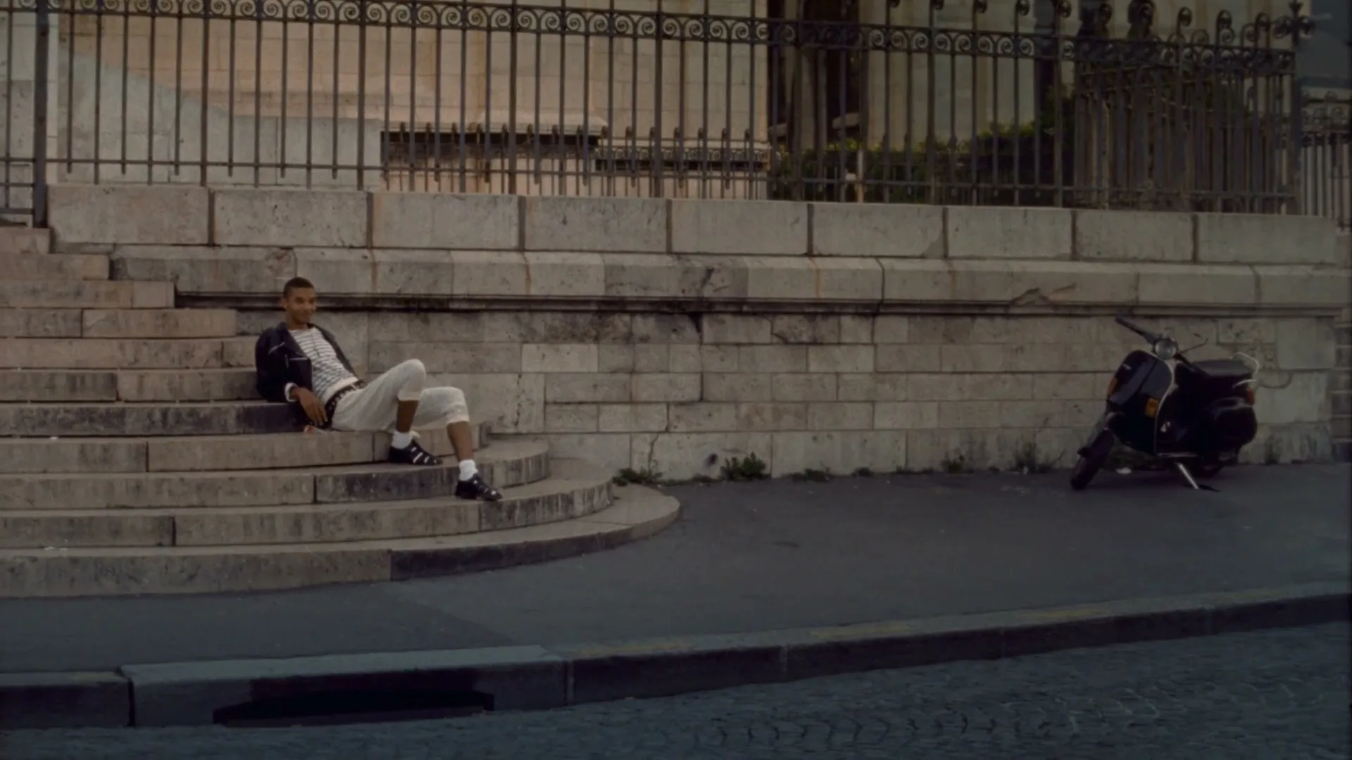 A young man in a striped jacket and white pants sits on stone steps next to a parked scooter on a street in Paris.