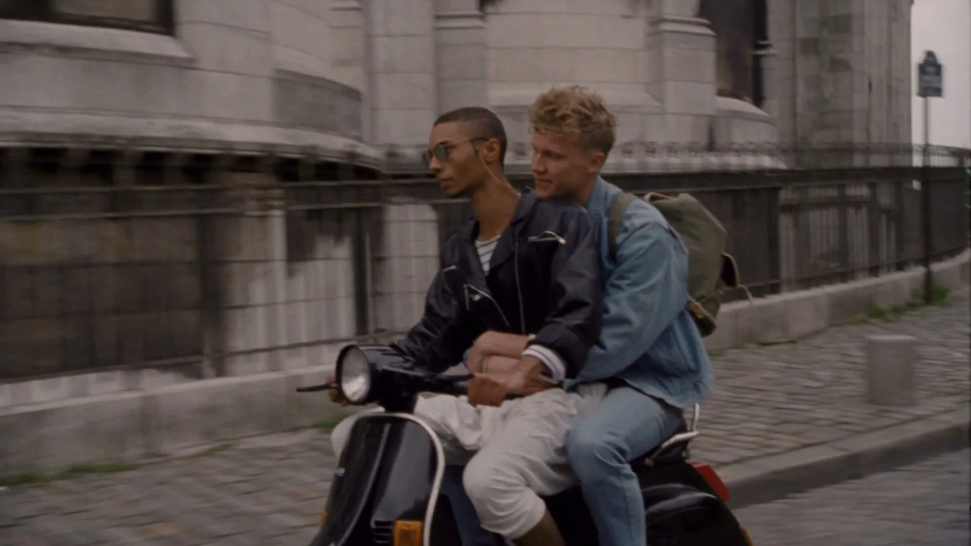 Two young men ride a black scooter on a cobblestone street in front of a stone building with a fence.