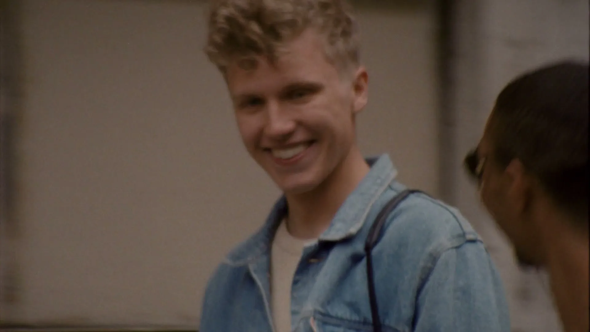 A young man with curly blonde hair in a denim jacket smiles while talking to another man, partially visible, on a city street.