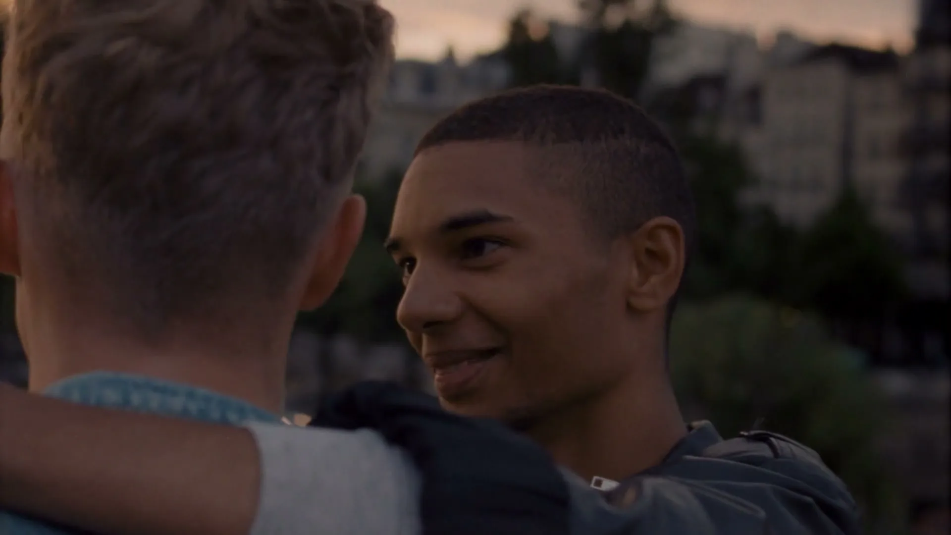 A young man with short dark hair smiles at another man, whose back is to the camera, as they embrace outdoors at dusk.