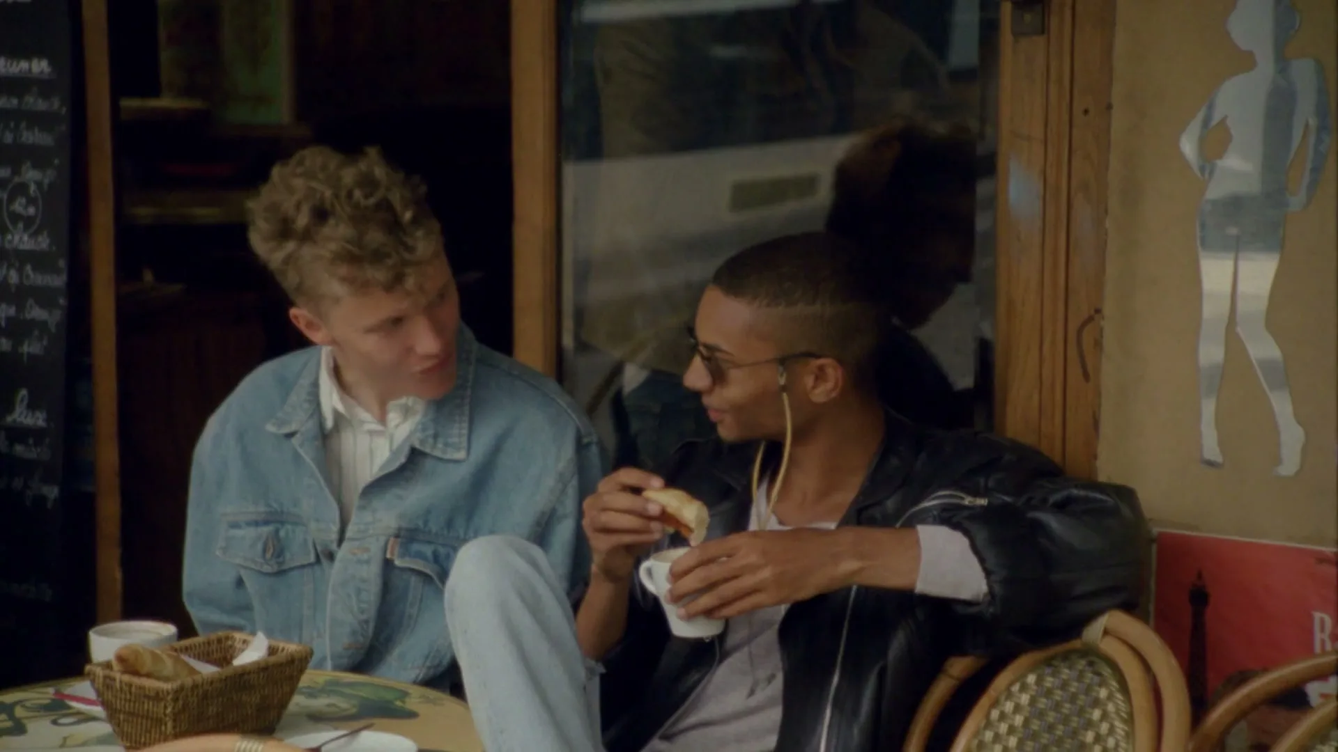 Two young men sit at an outdoor cafe table, one in a denim jacket and the other in a leather jacket, eating and talking.