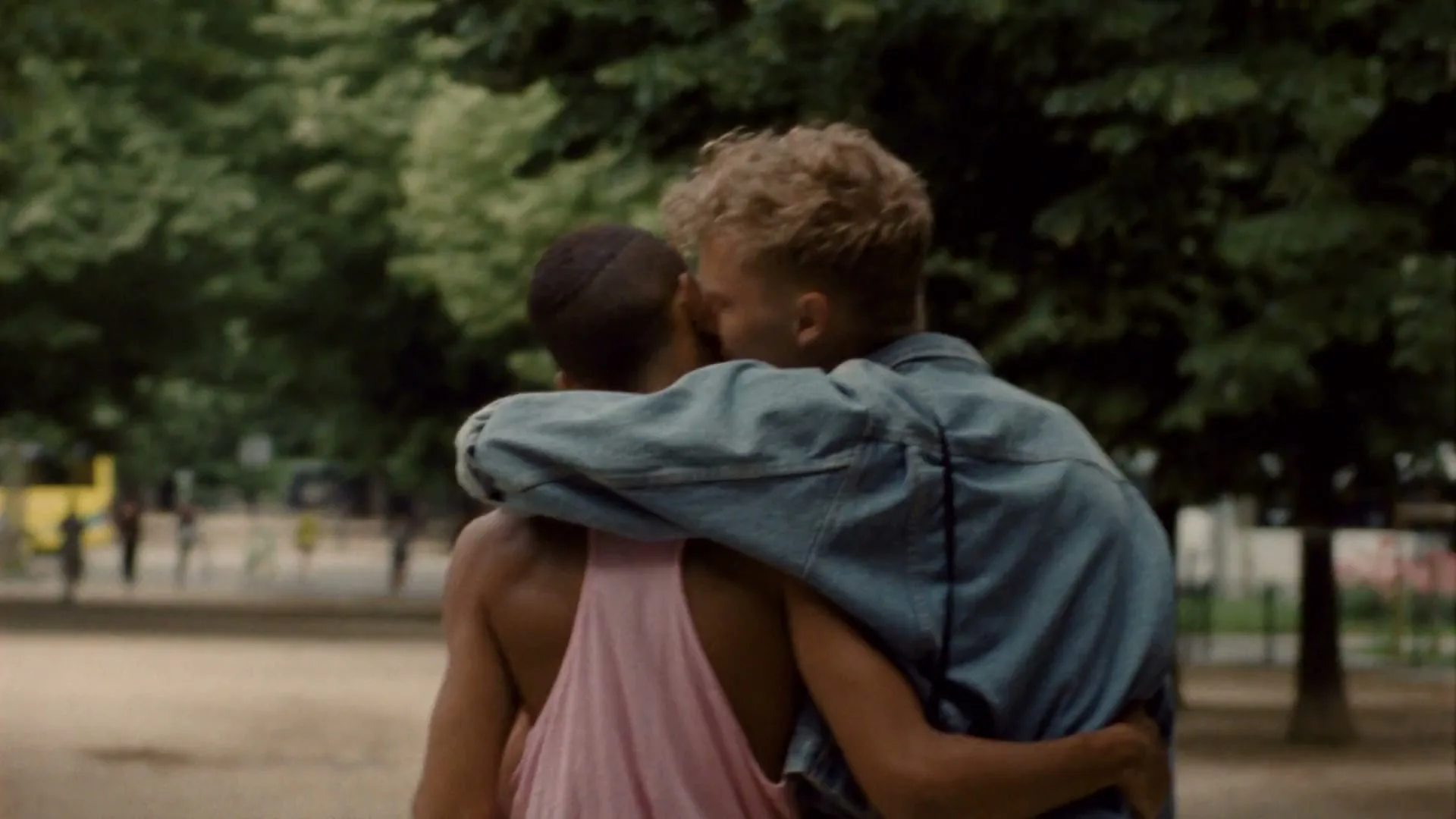Two young men, one with an arm around the other, walk away from the camera in a park with green trees in the background.