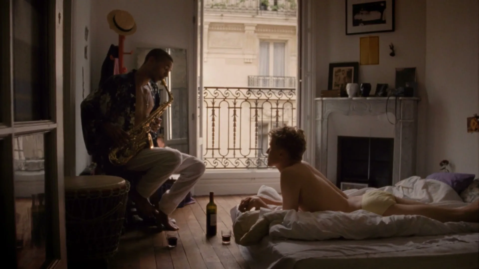 A man plays saxophone for a woman lying in bed in a Parisian apartment, with an open balcony door revealing the city outside.