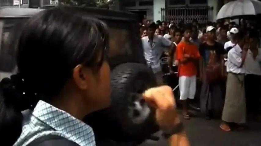 A young woman with dark hair in a ponytail, wearing a plaid shirt and backpack, raises her fist in protest as cars drive by, with a crowd of people watching in the background on a street.