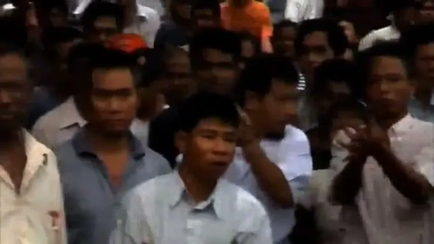 A crowd of people, mostly men, are gathered outdoors, some looking around and one man raising his hand.