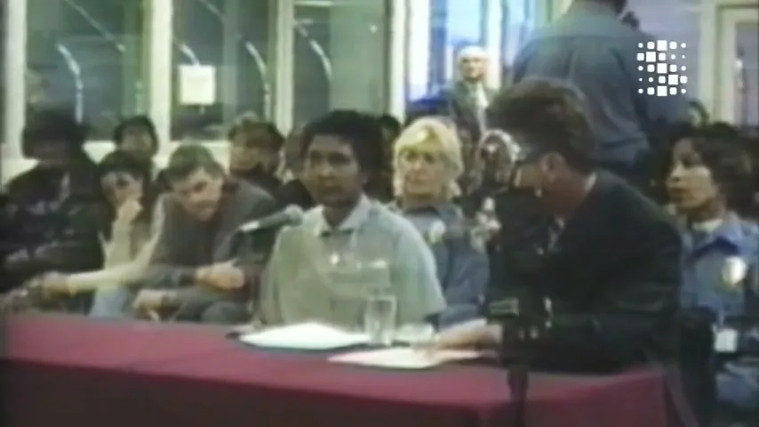 A woman with dark skin and short dark hair sits at a table with a microphone, speaking to a group of people in an interior setting.