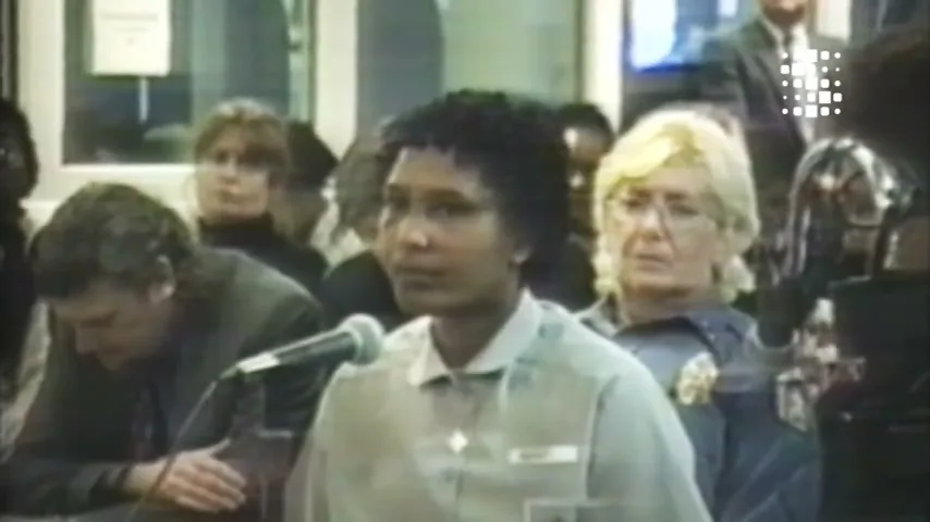 A woman with dark curly hair and a light-colored shirt speaks into a microphone in a courtroom, surrounded by other people.