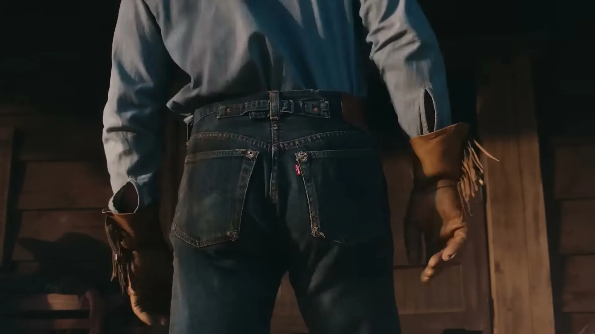 A person wearing blue jeans and a light blue shirt stands with their back to the camera, in a dimly lit wooden interior.