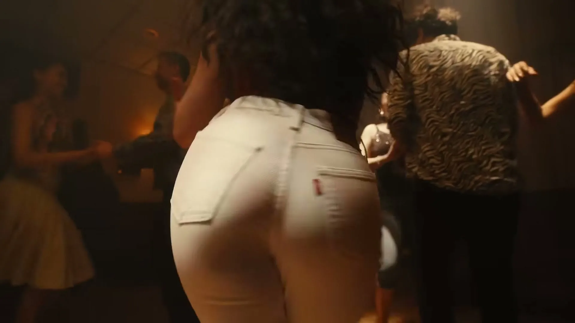 A woman in white jeans dances with her back to the camera, surrounded by other people dancing in a dimly lit room.
