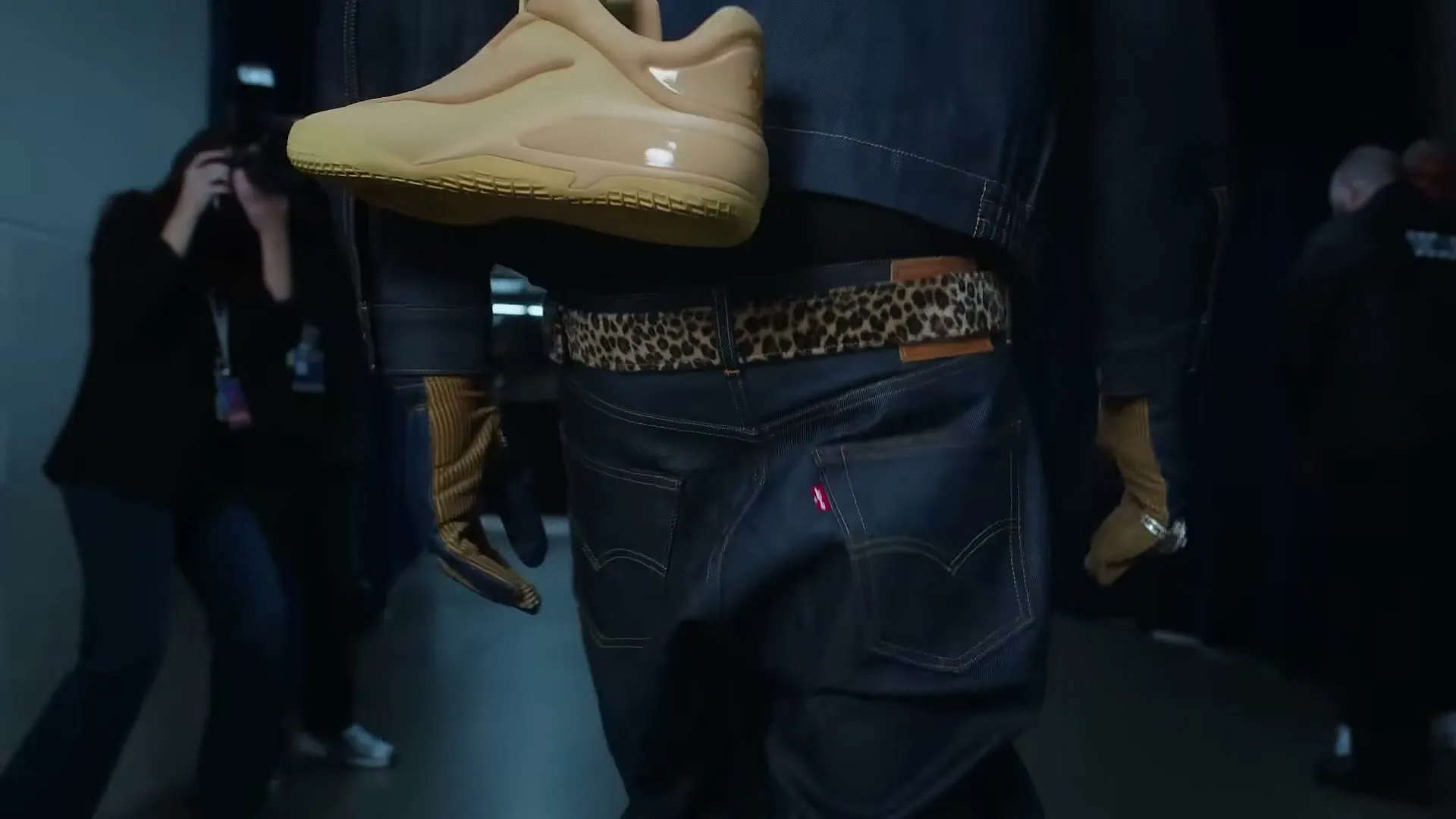 A person in a denim jacket and jeans with a leopard print belt walks away from the camera, holding a tan shoe, in a dark hallway.