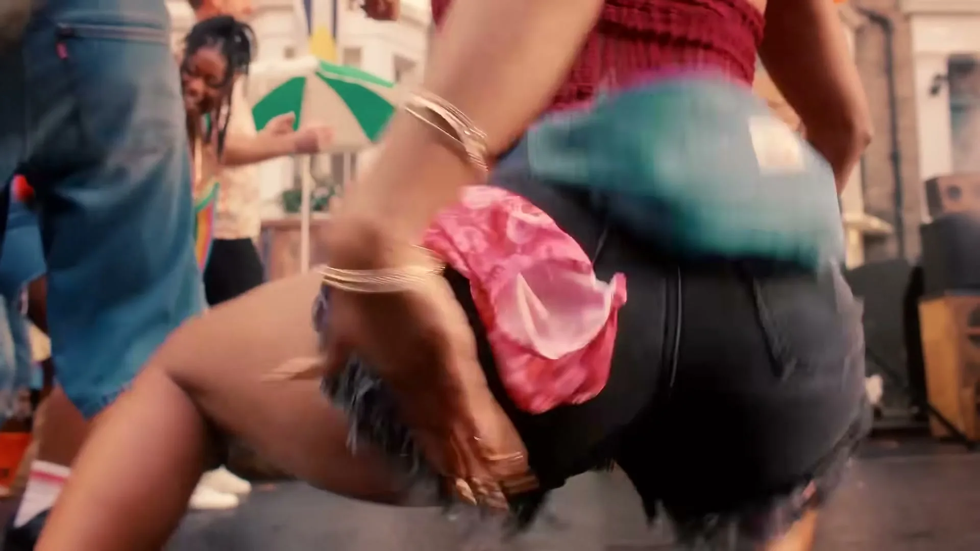 A person in black denim shorts and a red top dances with their back to the camera, with other people dancing in the background on a street.