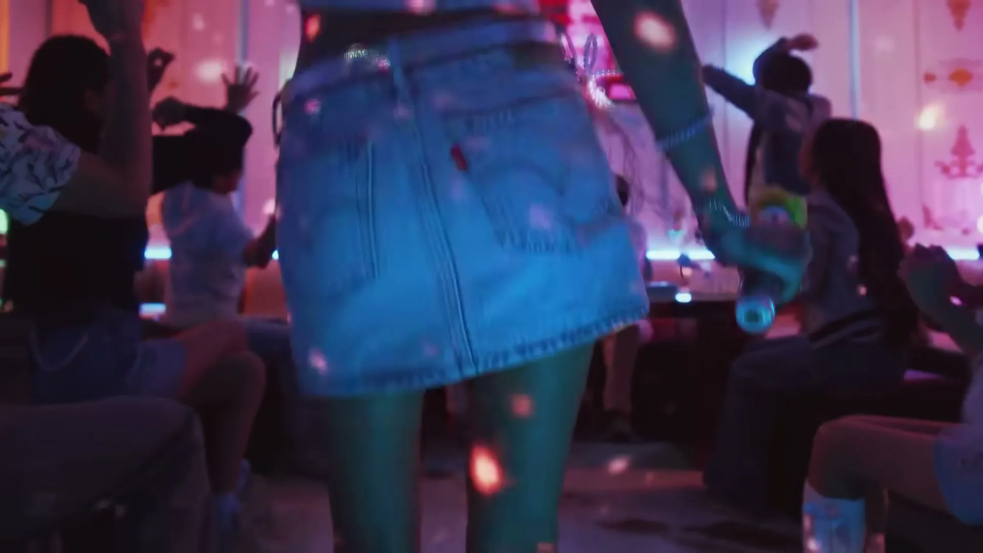 A young woman in a denim skirt dances in a crowded, neon-lit club, with disco lights reflecting on her back.