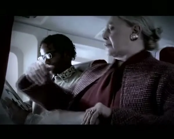 A woman in a red jacket and a man in glasses sit next to each other on an airplane, looking at a magazine.