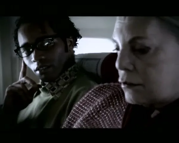 A young man in glasses and an older woman sit side-by-side on an airplane, looking forward as the cabin darkens.