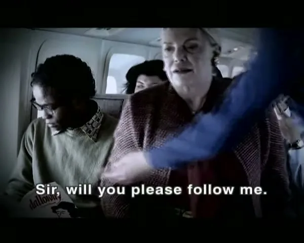 A flight attendant asks a woman to follow her, while a man sitting next to the woman smiles, on an airplane.