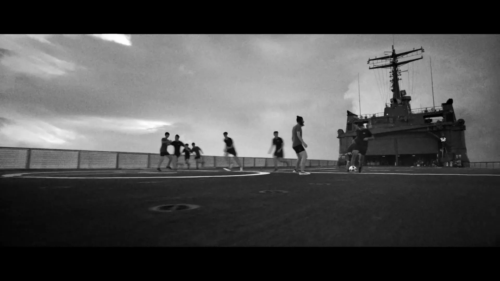 A group of male sailors in dark t-shirts and shorts play soccer on the deck of a large naval ship under a cloudy sky.