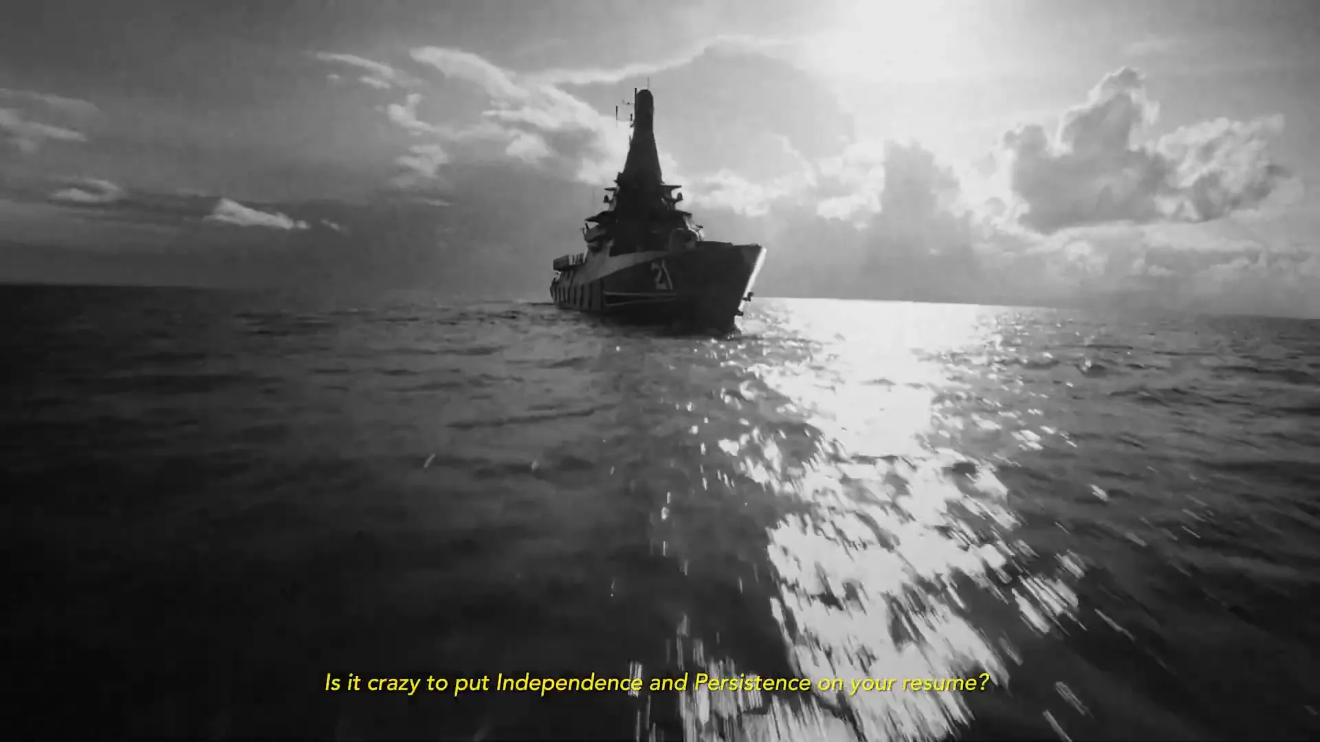 A warship with the number 21 on its bow sails on the open sea, with the sun reflecting on the water, in a black and white shot.