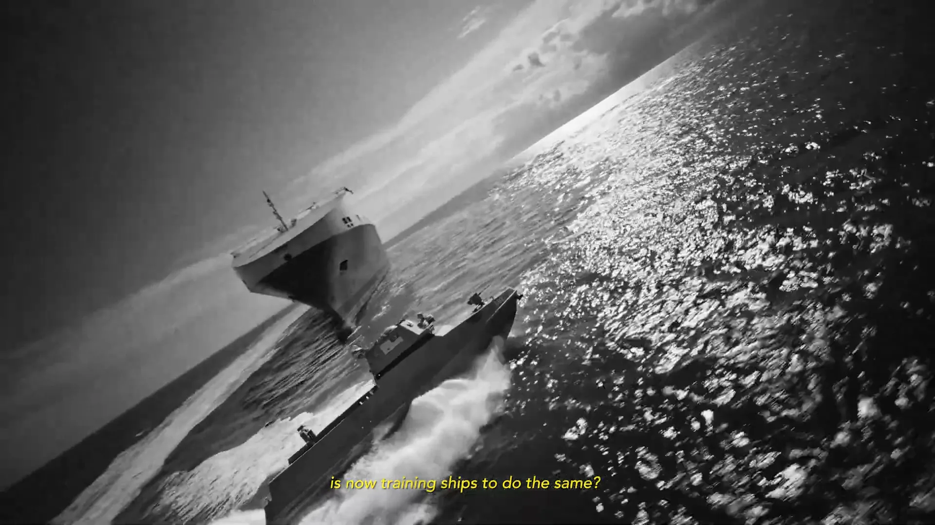 A black and white shot shows a large cargo ship and a smaller naval vessel sailing on a sparkling sea under a cloudy sky, with a hand holding tongs appearing from the top.