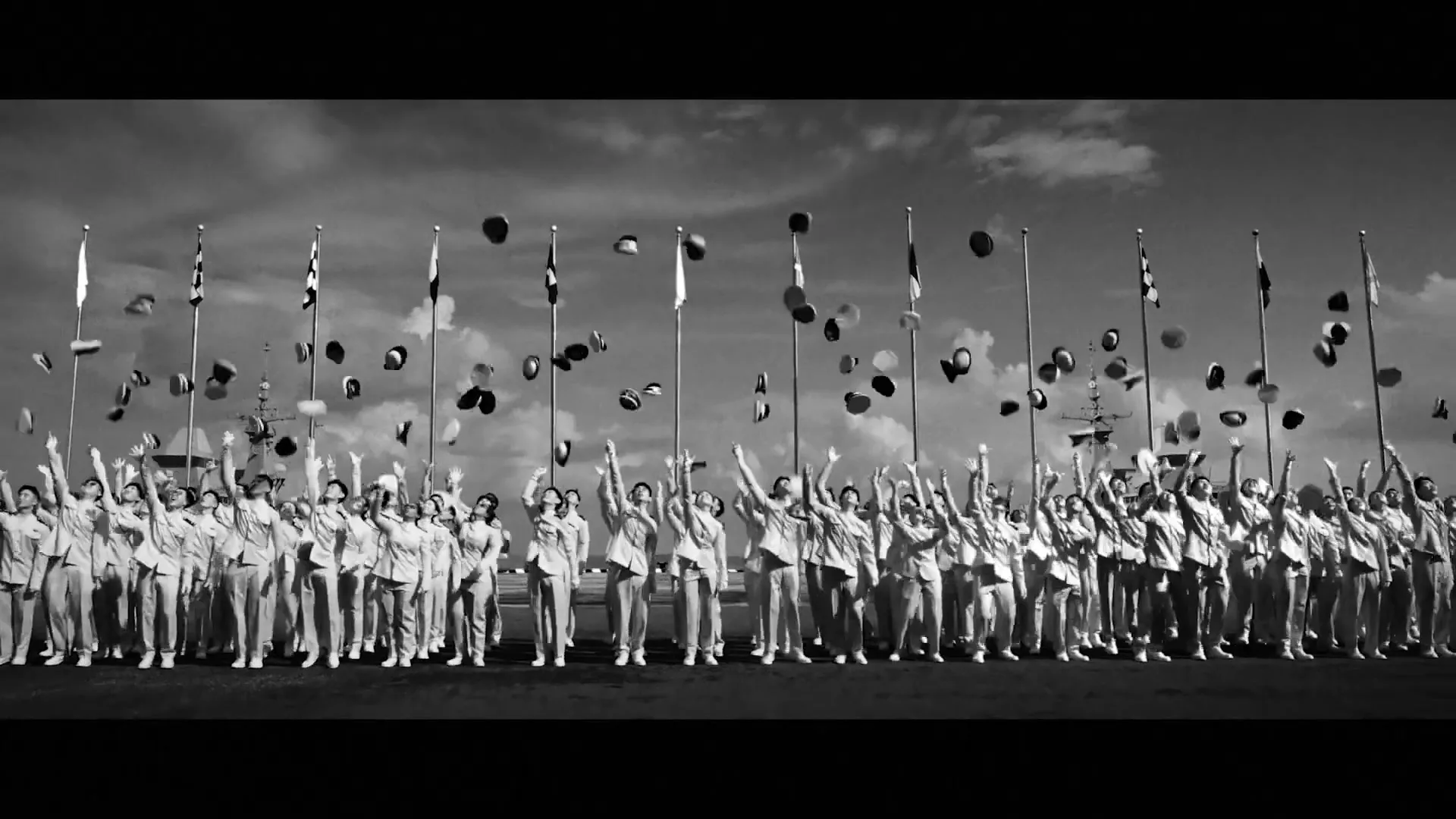A large group of diverse naval cadets in uniform throw their caps in the air in celebration, standing in front of flagpoles and warships under a cloudy sky.