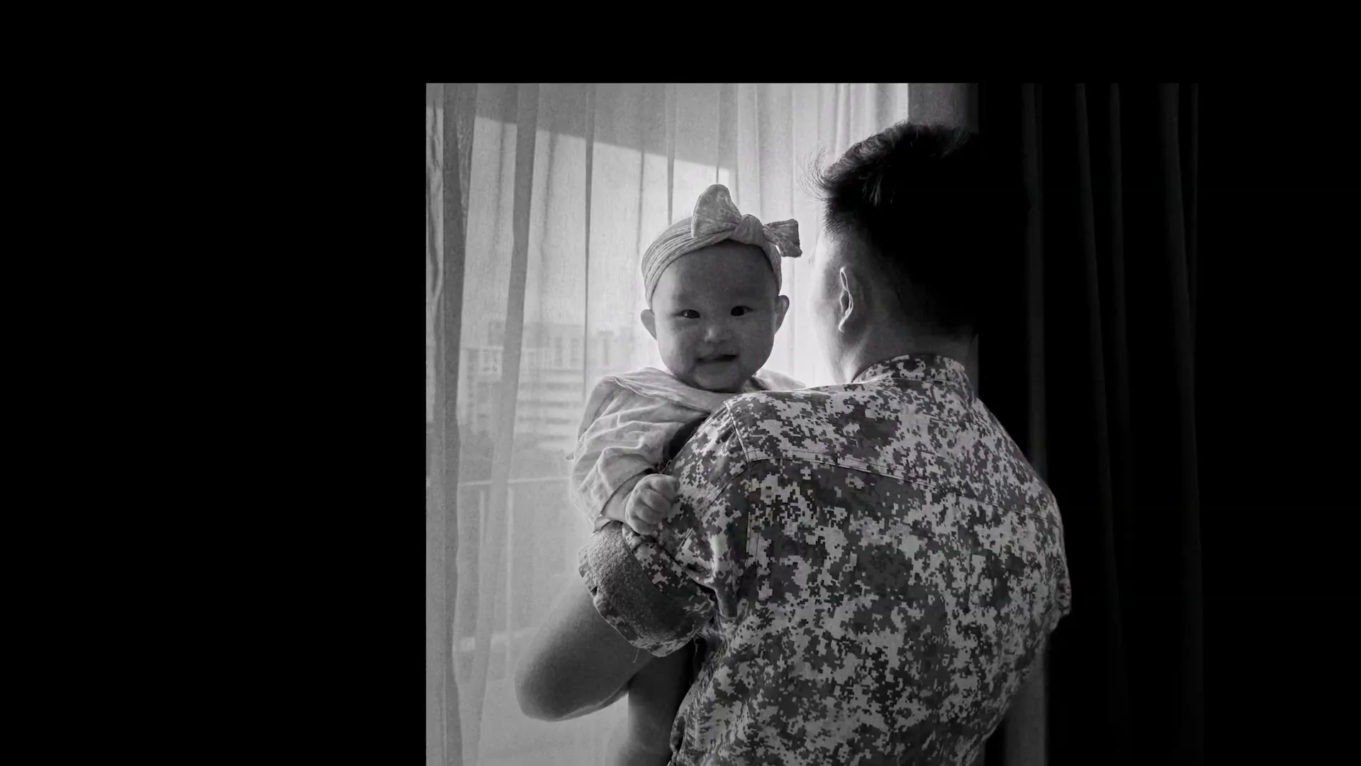 A man in a camouflage shirt holds a baby wearing a bow headband, looking at the camera in a room with a window.