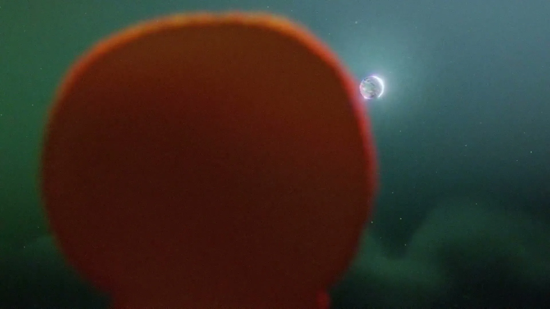 A blurry, out-of-focus orange sphere dominates the foreground, while a small, glowing planet-like object with a crescent shape is visible in the distant, dark green background.
