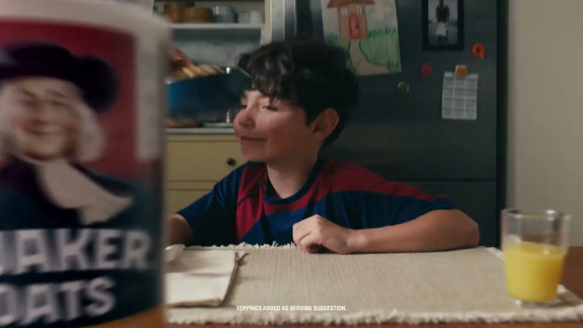 A young boy with curly hair sits at a kitchen table, looking up as a woman's hands place a bowl of oatmeal in front of him, with a Quaker Oats container in the foreground.