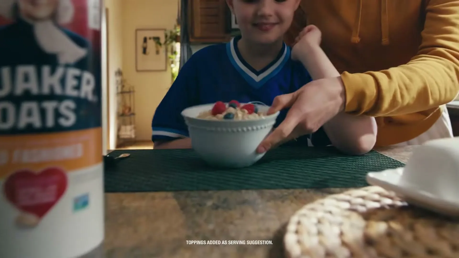 A child in a blue soccer jersey smiles as a bowl of oatmeal with berries is placed in front of them on a kitchen counter, with a Quaker Oats container in the foreground.