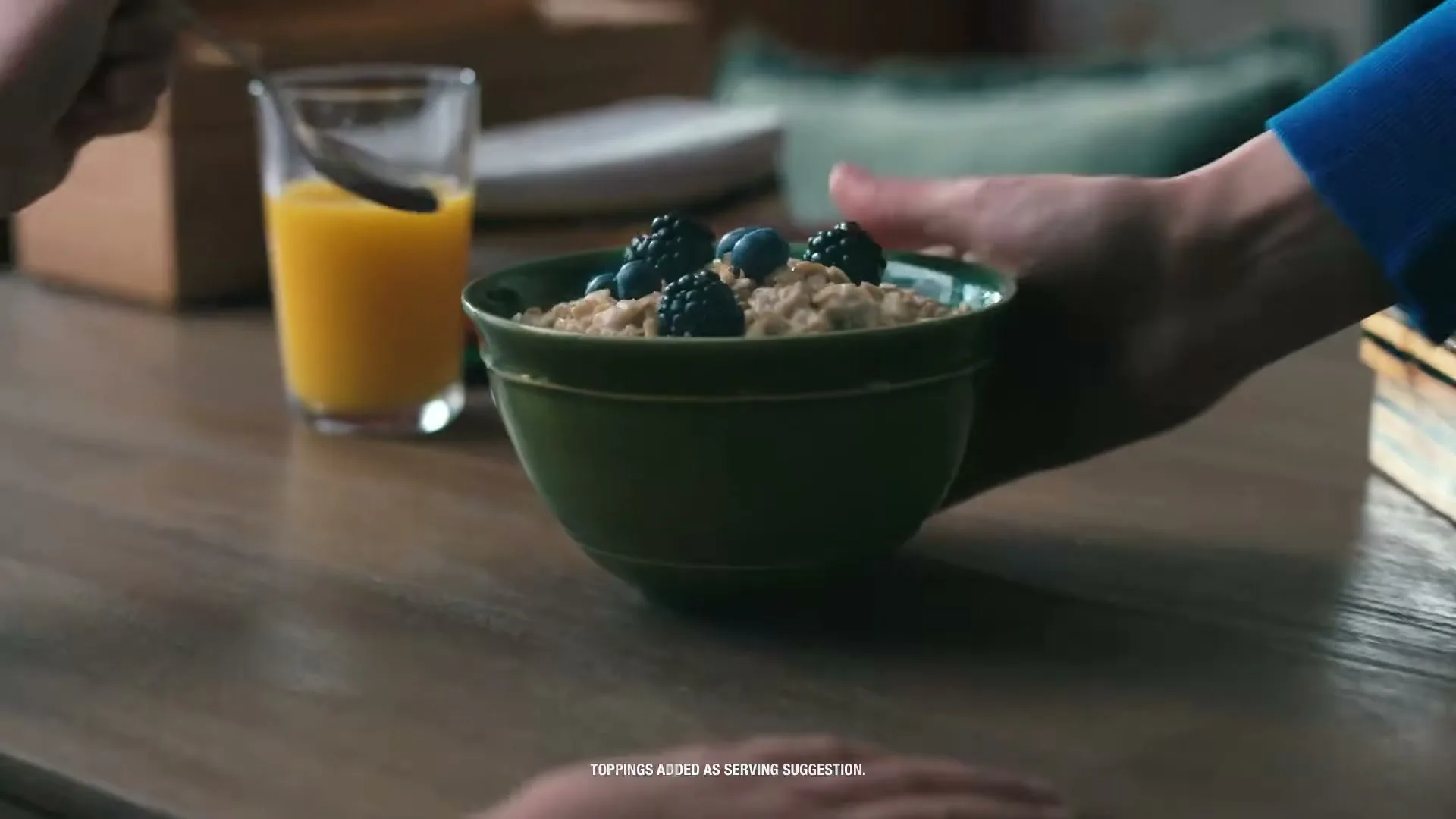 A person's hands place a green bowl of oatmeal with berries and a glass of orange juice on a wooden table, with another person's hands visible in the foreground.