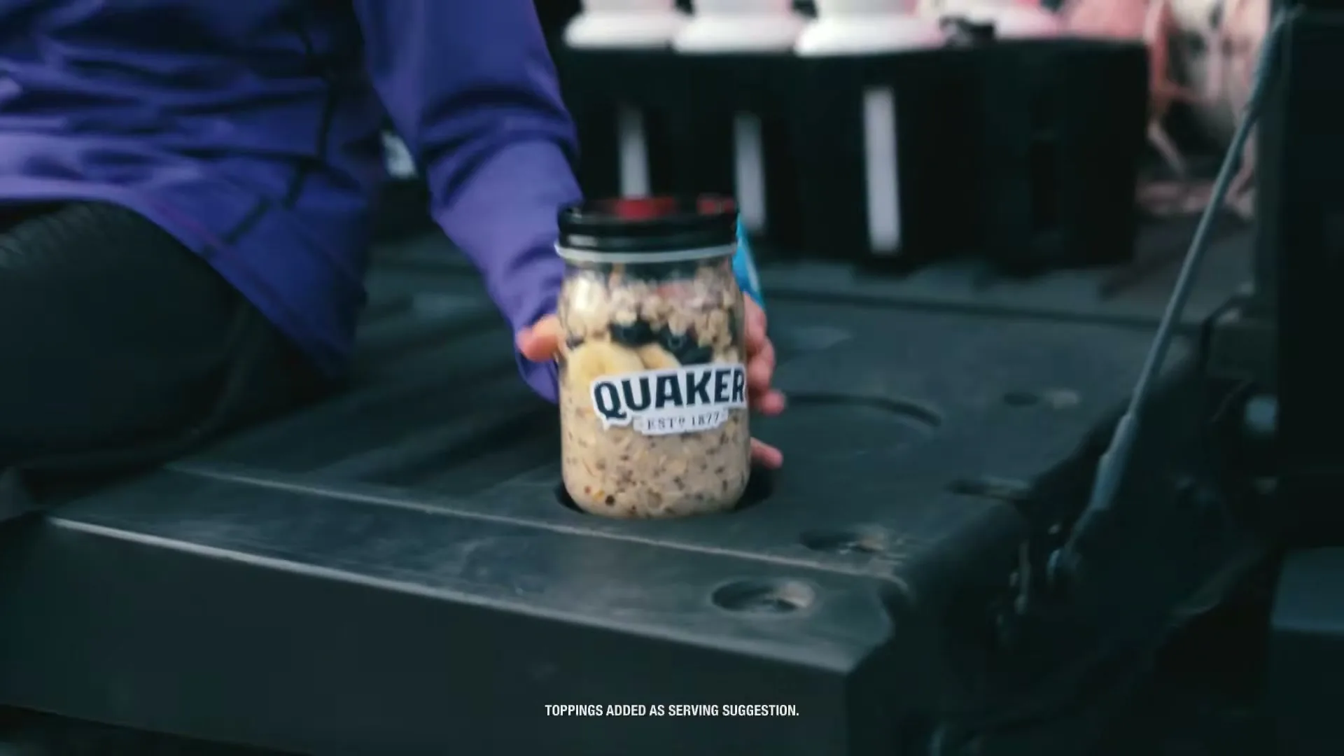 A person in a purple shirt places a jar of Quaker oatmeal with fruit on the tailgate of a truck.