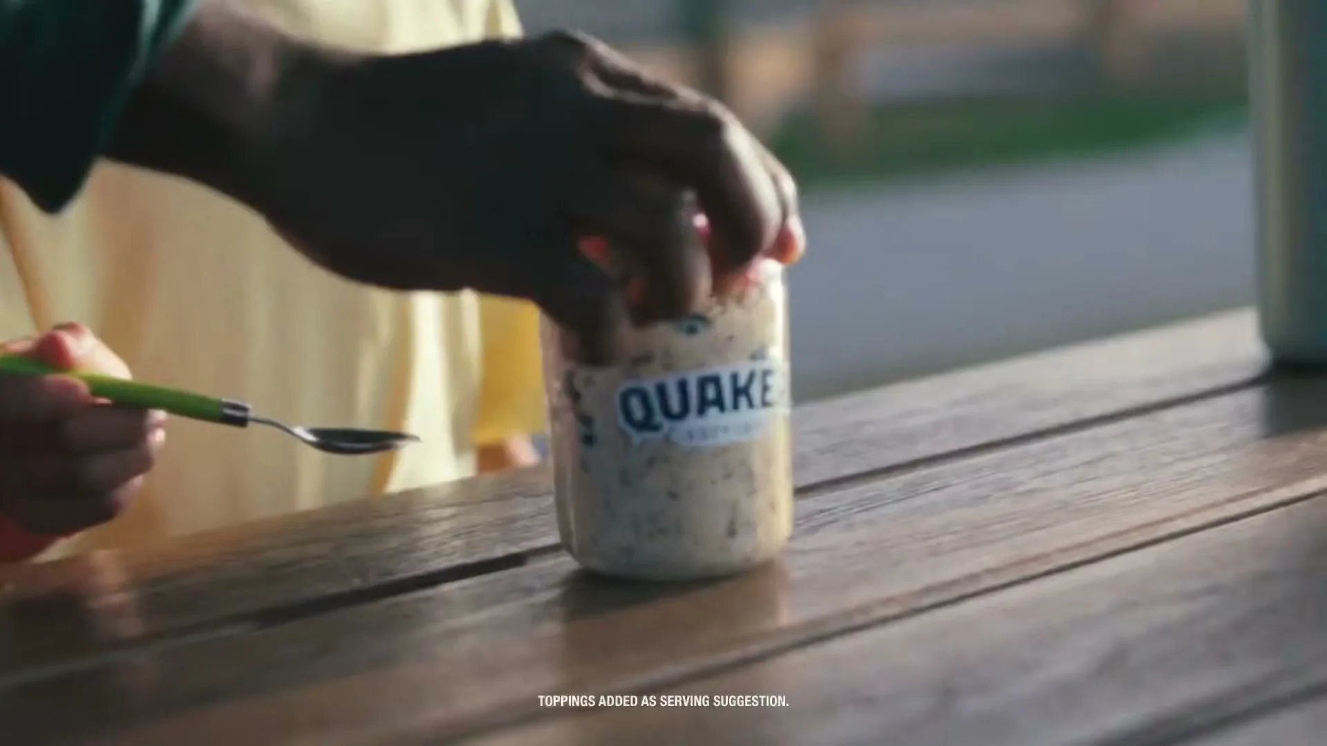 A person's hands open a jar of Quaker oats with berries on top, on a wooden table outdoors.