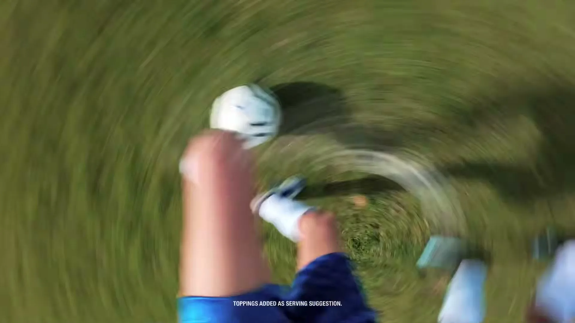 A person's legs and feet in blue shorts and white shoes kick a white soccer ball on a green grass field from a high angle.