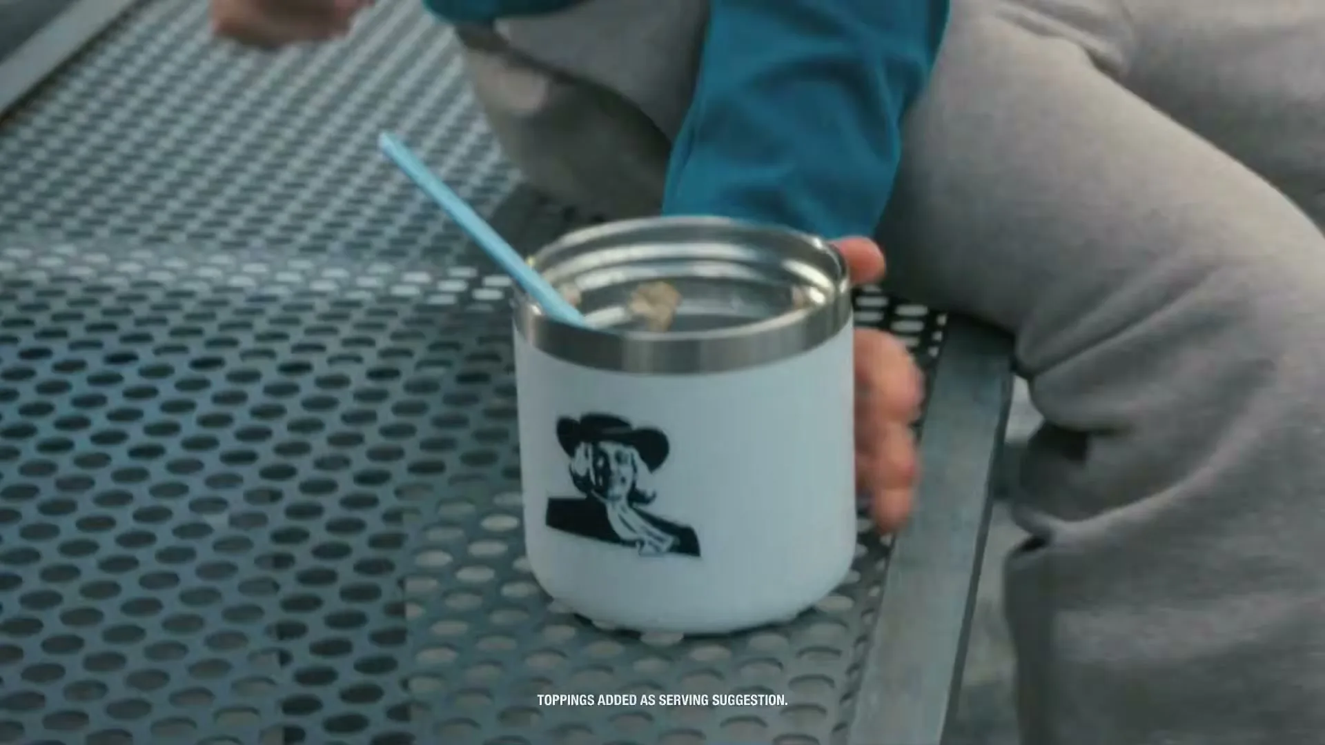 A person in grey sweatpants and a blue jacket places a white insulated cup with the Quaker Oats logo and a blue straw on a metal bench.