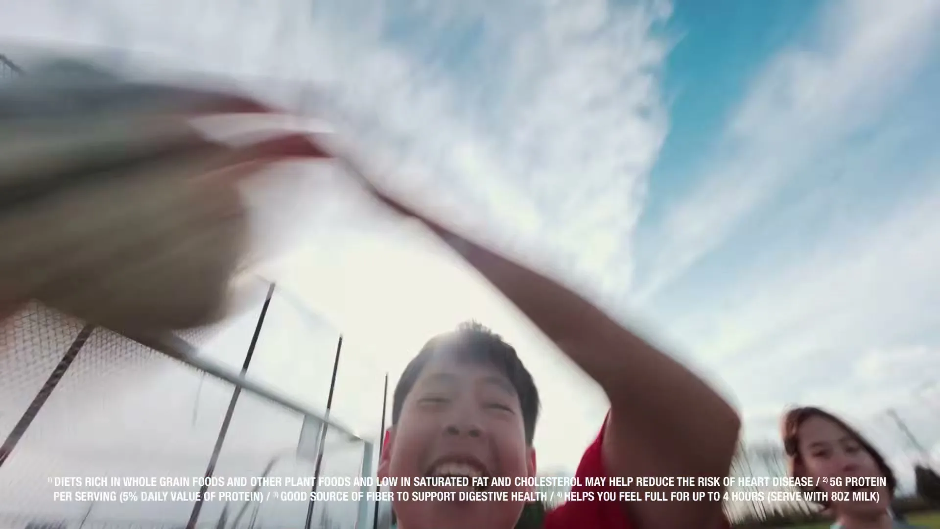 A young boy with dark hair and a red shirt looks up and smiles, with a soccer ball flying above him against a blue sky.