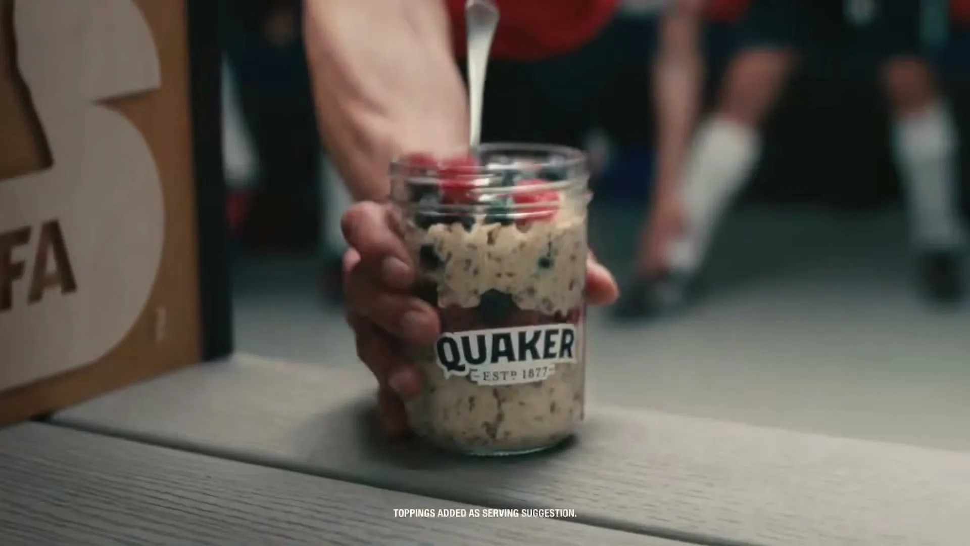 A person's hand places a jar of Quaker oatmeal with berries on a bench in a locker room, with soccer players in the background.