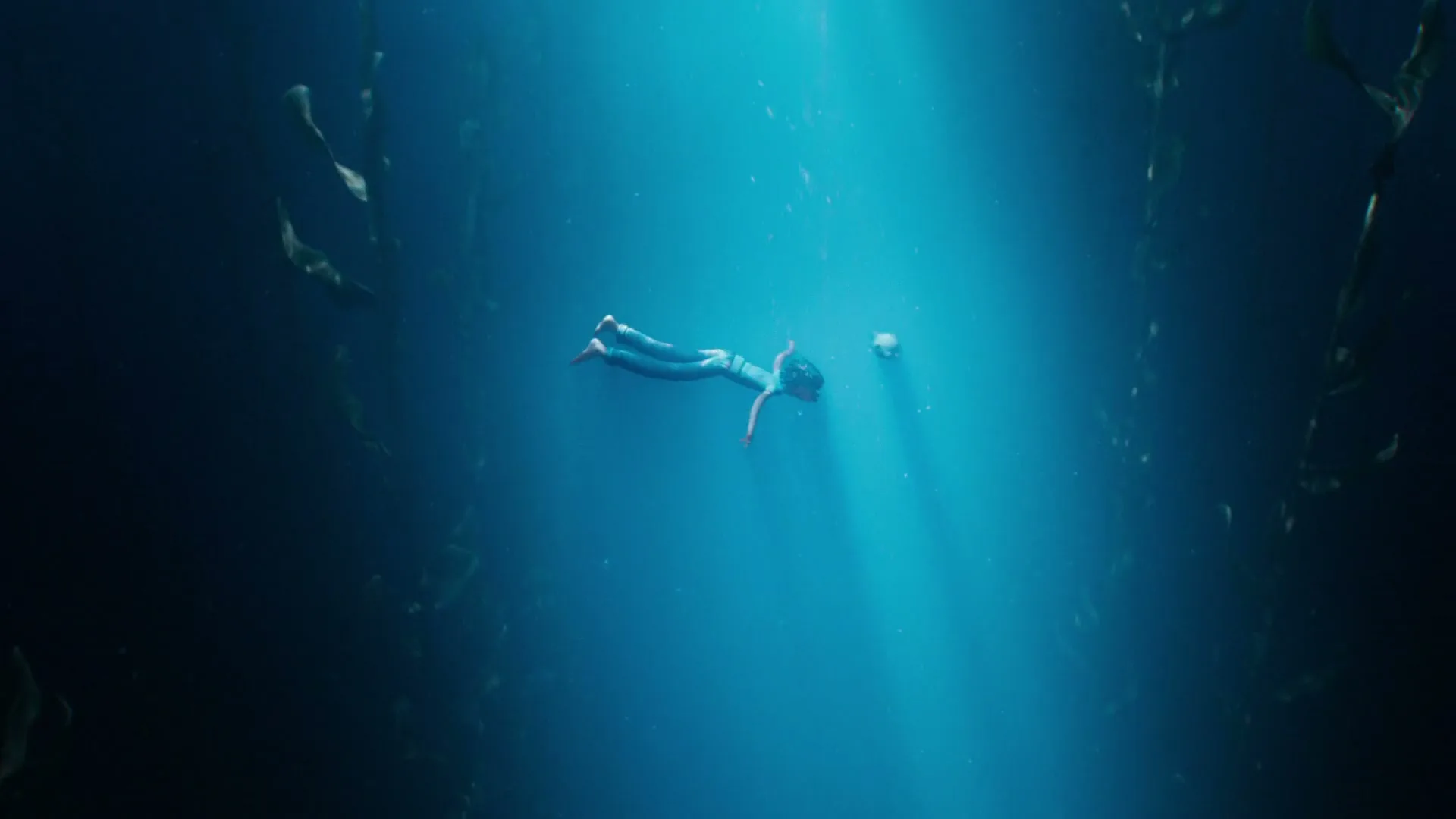 A young woman in a wetsuit swims underwater with a glowing device, surrounded by kelp, in a deep blue ocean.