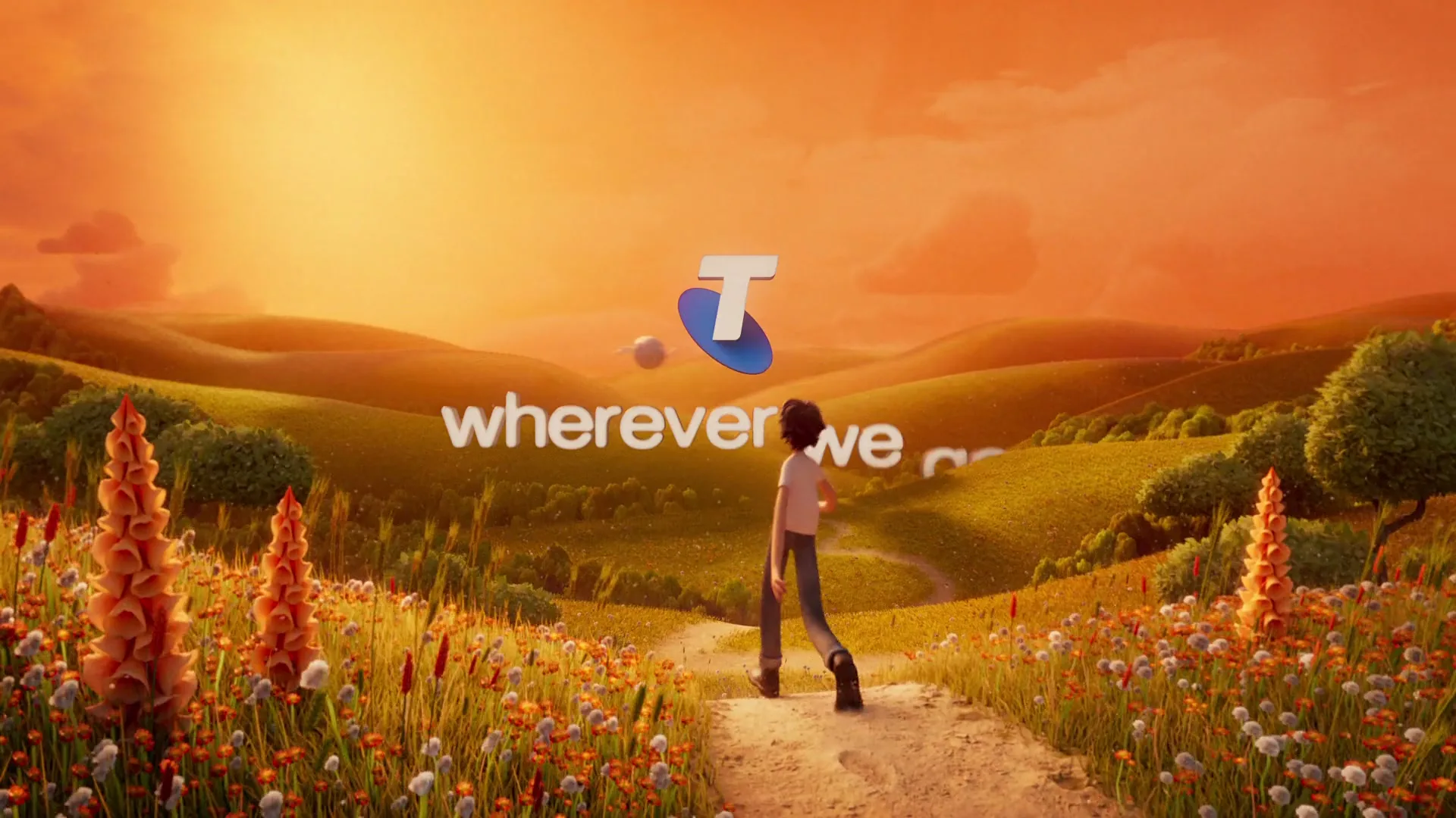 A young boy walks along a dirt path through a field of wildflowers at sunset, with the Telstra logo and tagline appearing in the sky.