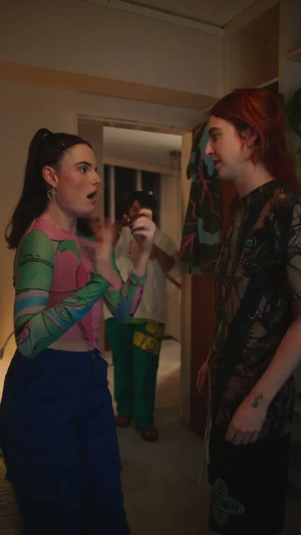 Two young women stand in a doorway, talking animatedly, while a third person is visible in the background, partially obscured.