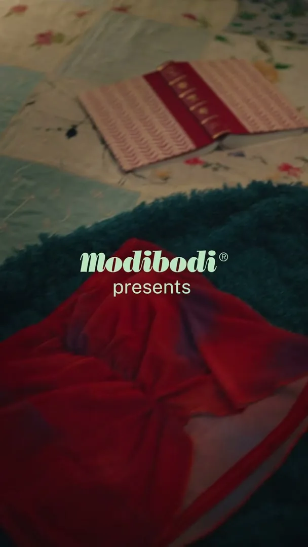 A pair of red underwear is placed on a teal blanket, with a patterned quilt and a book in the background, as a hand reaches in to cover the underwear with a black cloth.