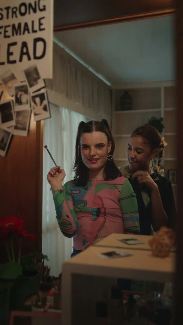 Two young women are in a bedroom, one applying makeup in a mirror while the other braids her hair, with a "STRONG FEMALE LEAD" sign visible.