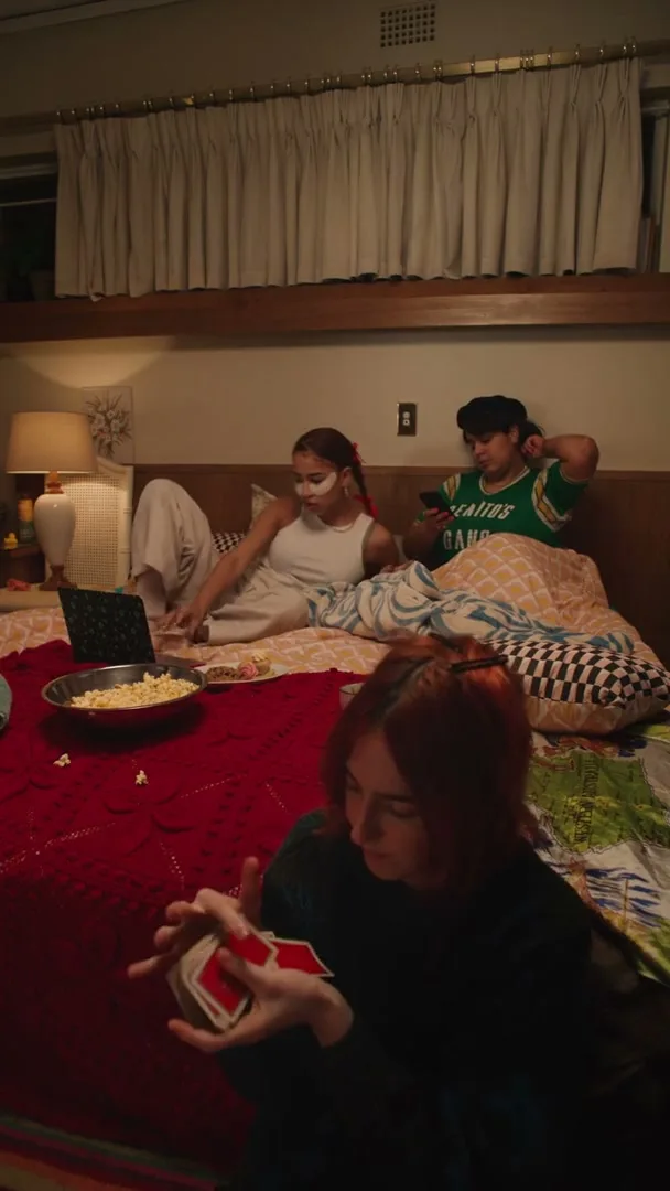 Three young women are having a sleepover in a bedroom, one shuffles cards in the foreground while two others relax on the bed behind her.