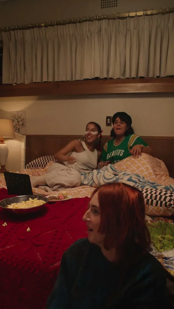 Three young women are having a sleepover, watching something on a laptop, eating popcorn, and laughing in a cozy bedroom.