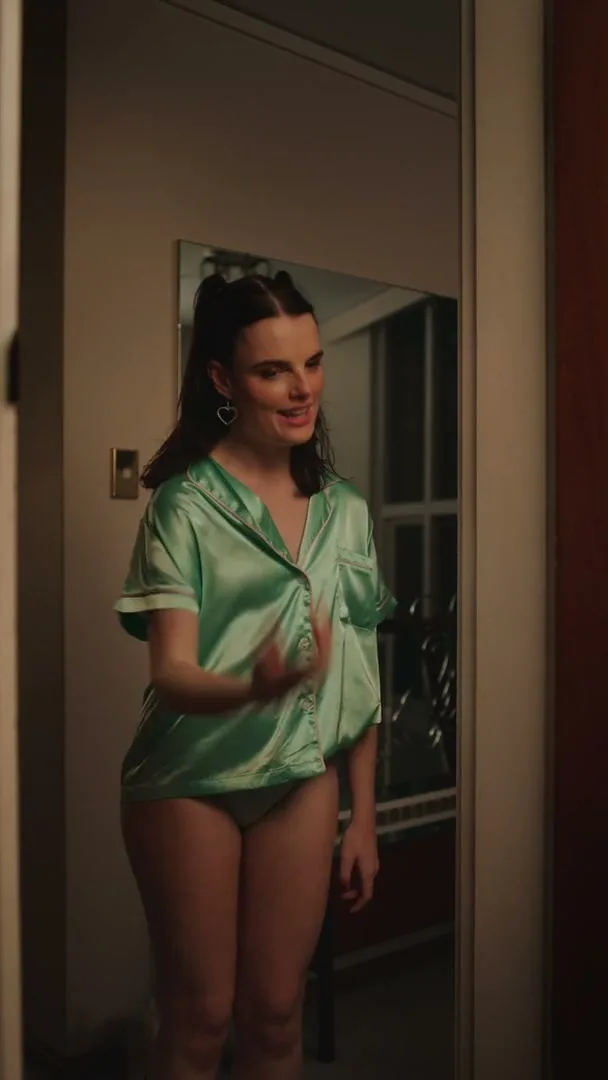 A young woman in a green silk pajama top and underwear stands in a doorway, looking around and talking to someone off-screen in an interior setting.