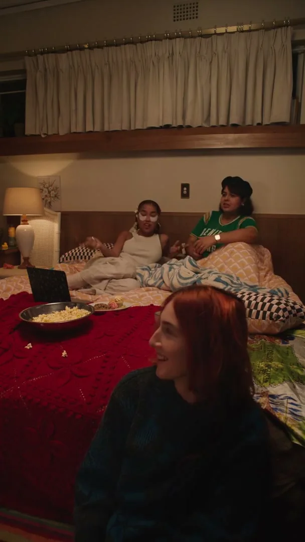 Three young women are having a sleepover in a bedroom, with two sitting on the bed and one on the floor, watching something on a laptop and eating popcorn.