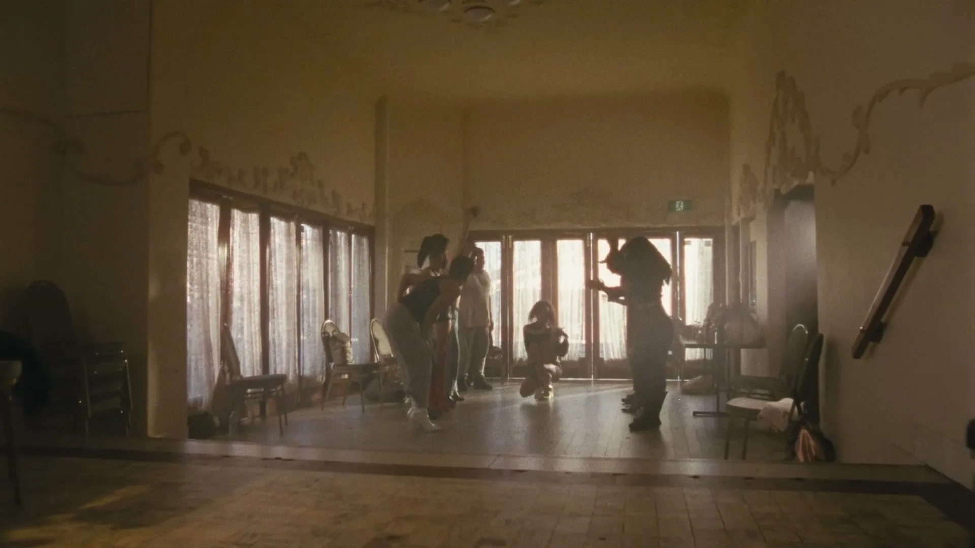 A diverse group of people dance energetically in a sunlit, somewhat dilapidated hall with large windows and ornate wall decorations.