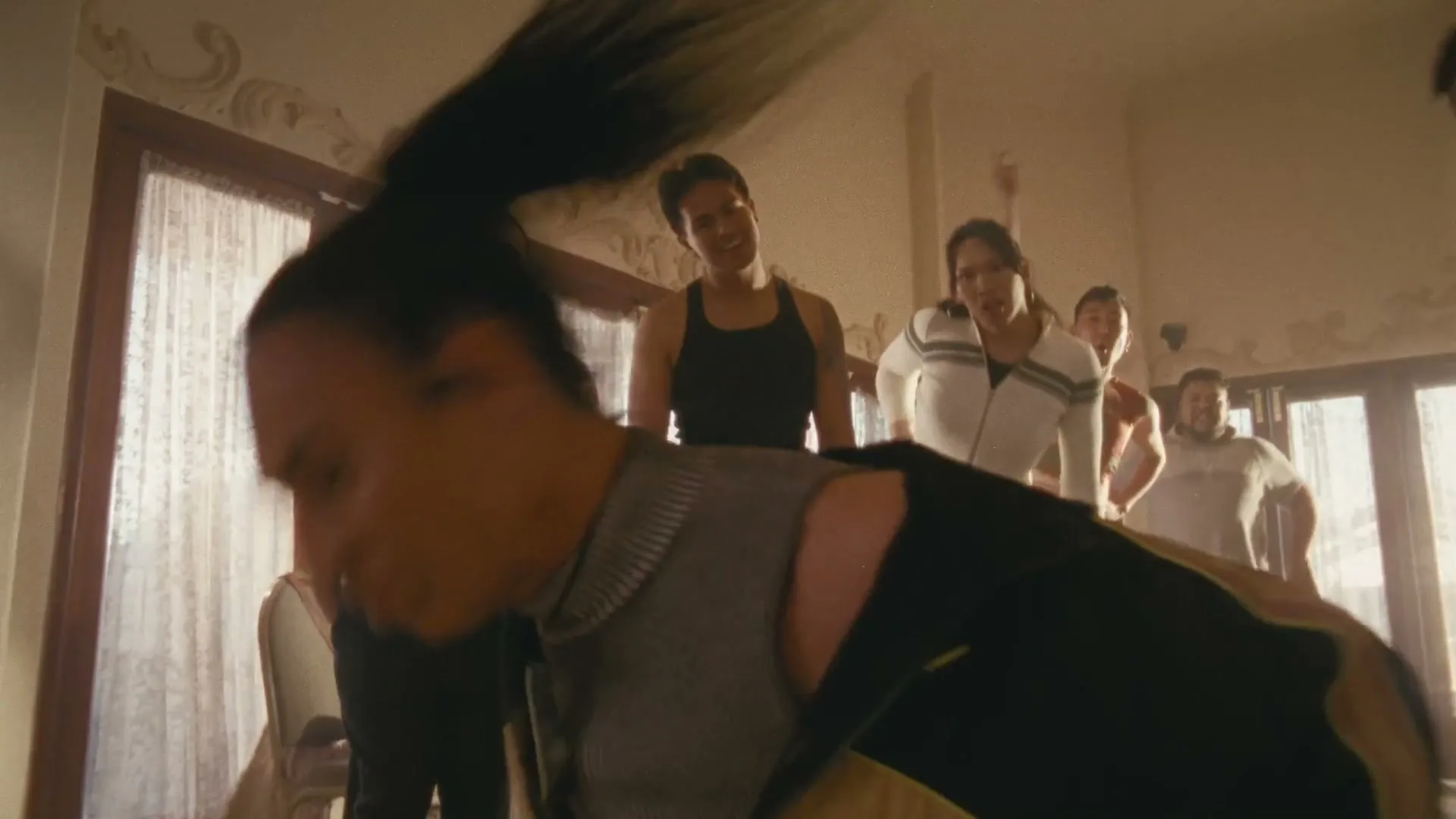A diverse group of people in athletic wear dance energetically in a sunlit room, with one person in the foreground bending over.