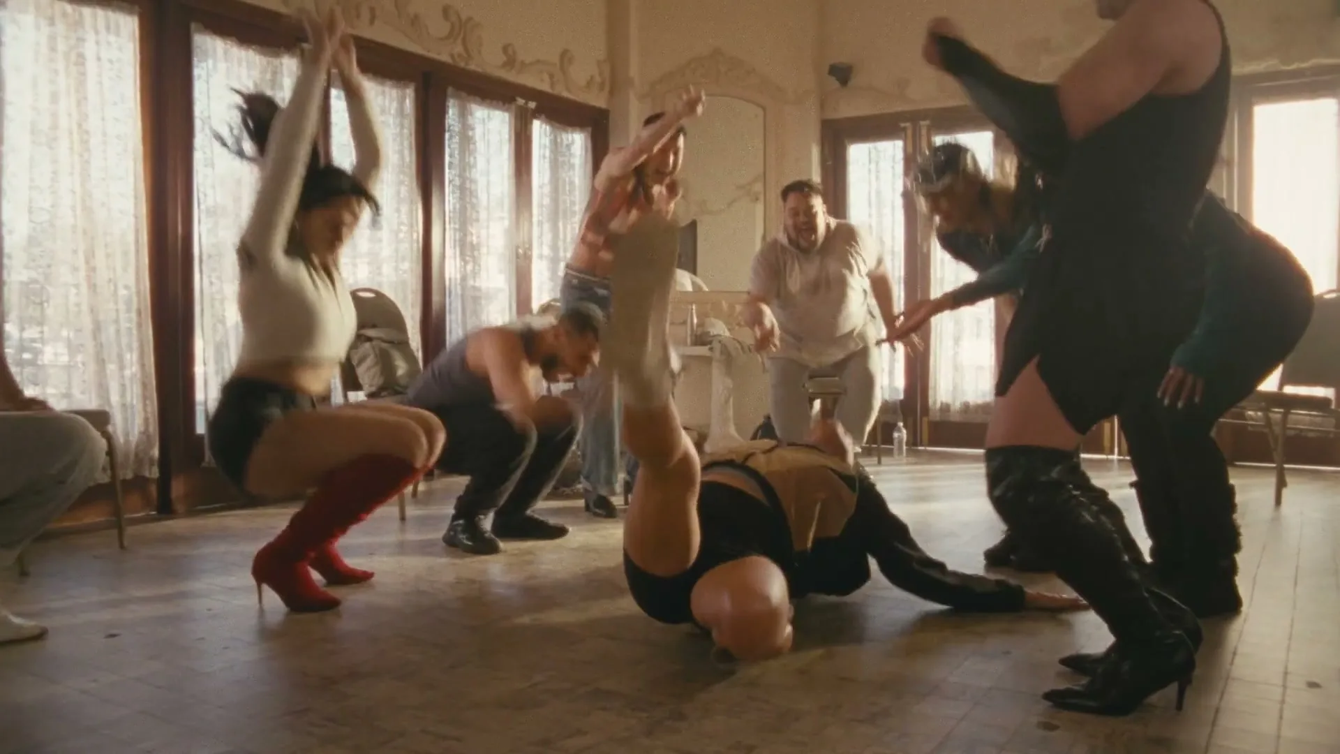 A diverse group of people in various outfits dance energetically in a sunlit room with large windows and white curtains.