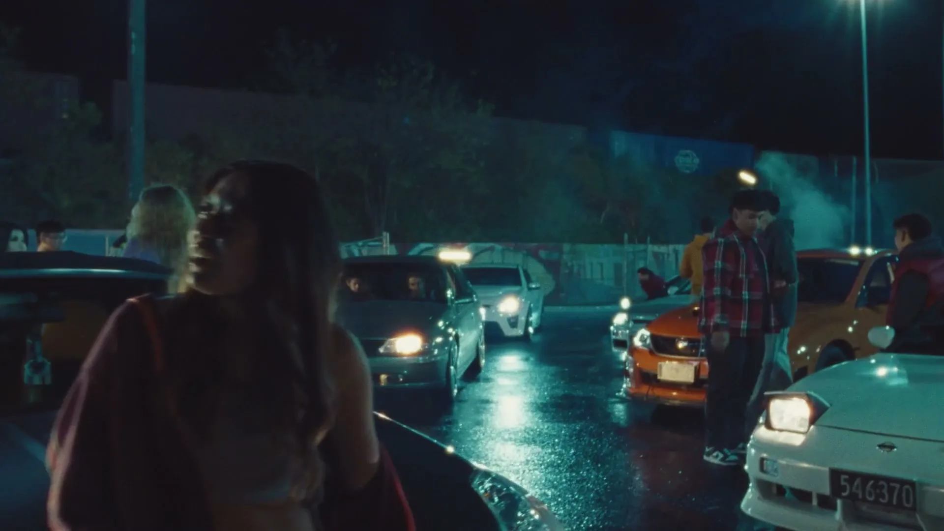 A young woman in a red jacket smiles and looks around at a night street racing event, with many cars and people in the background.